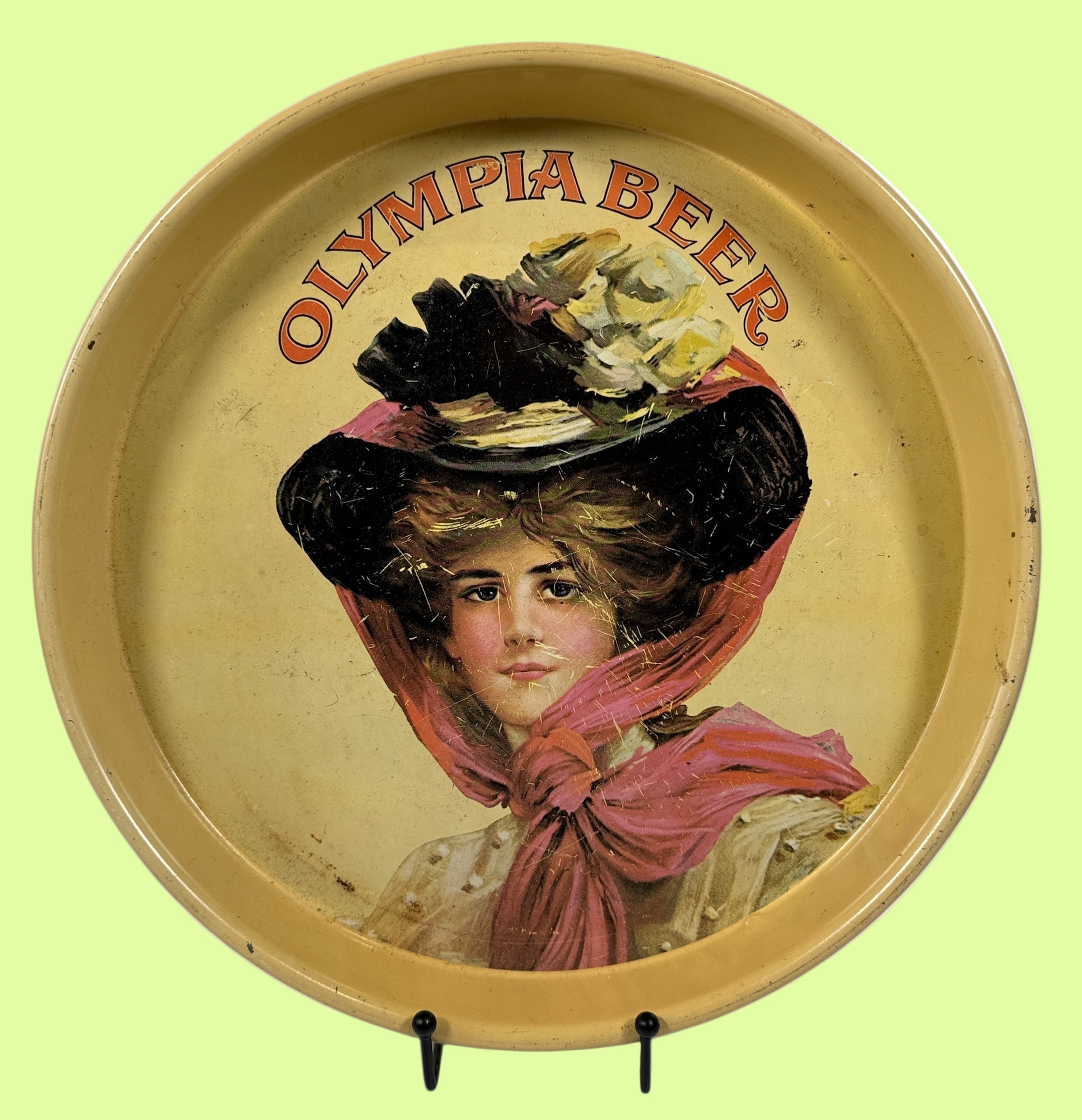 Round tray with a vintage illustration of a woman wearing a large hat and scarf, with 'Olympia Beer' text above.