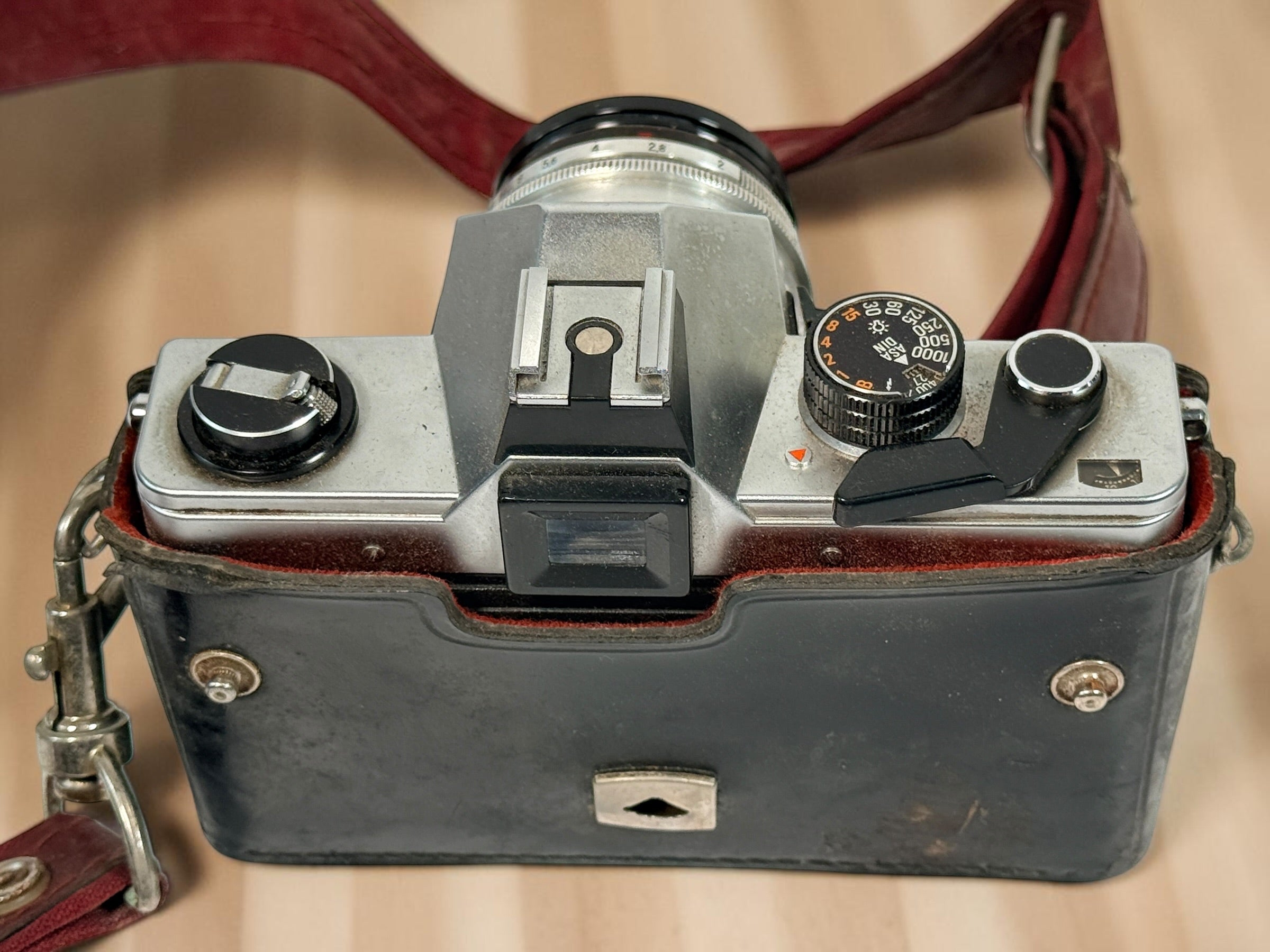 A vintage silver rangefinder camera housed in a black leather case with a reddish-brown leather strap, photographed from above on a light wood surface.