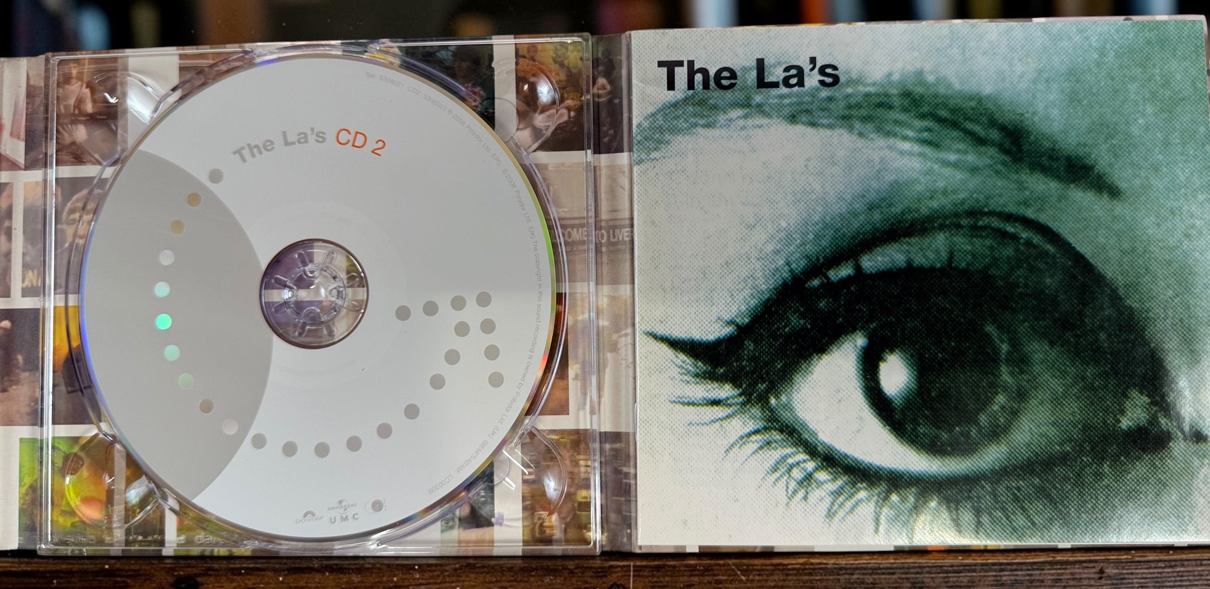 A CD case open to show the CD disc on the left and the album cover on the right, featuring the title 'The La's' and a large illustrated eye. The product is photographed on a wooden surface.