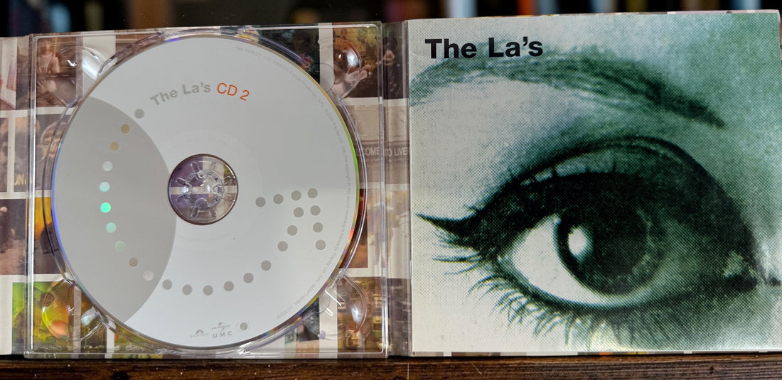 A CD case open to show the CD disc on the left and the album cover on the right, featuring the title 'The La's' and a large illustrated eye. The product is photographed on a wooden surface.