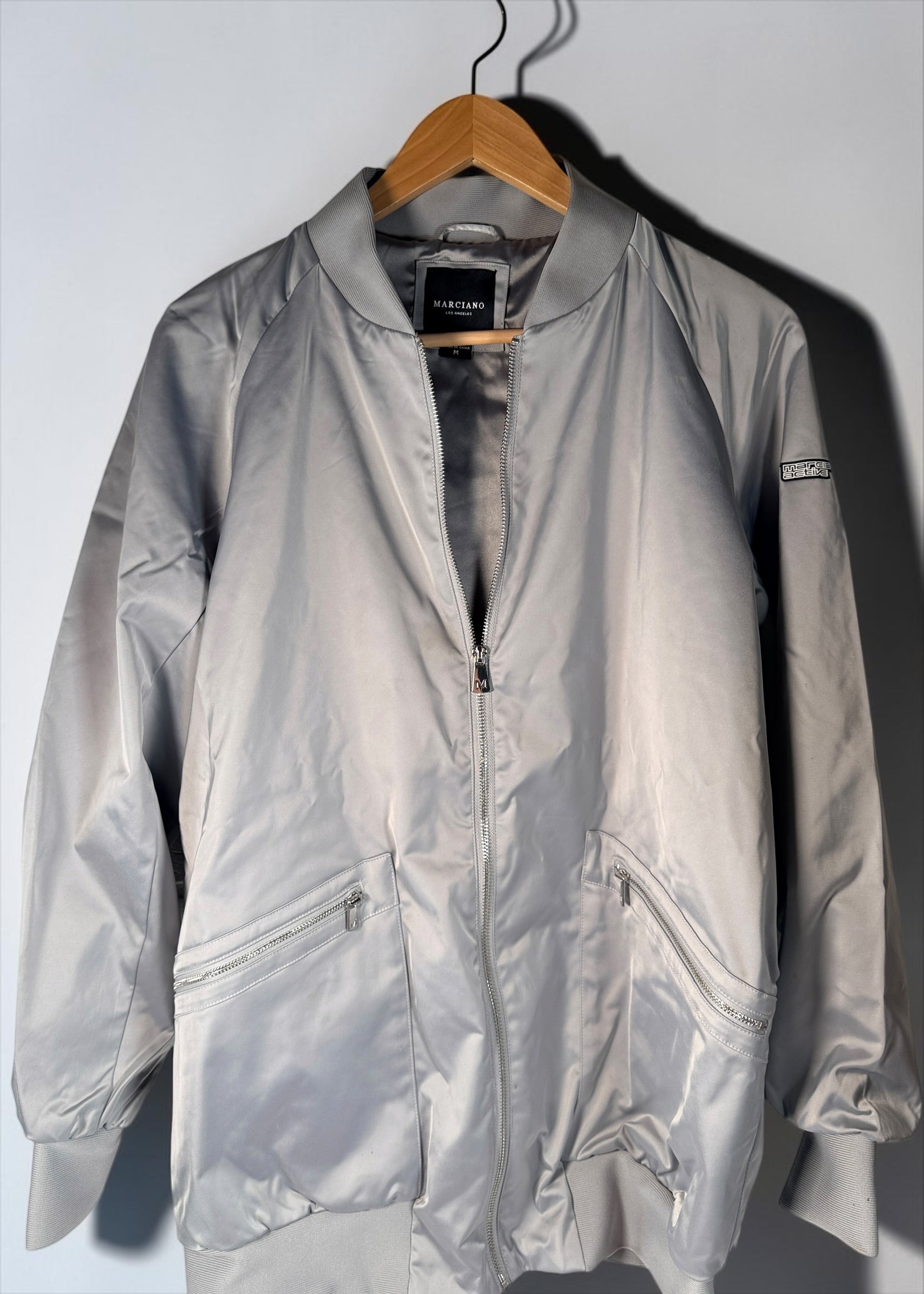 Shop the Marciano Limitless bomber jacket in silver-grey satin, size M. Original tags attached, zip pockets and ribbed trim. Minor marks disclosed.