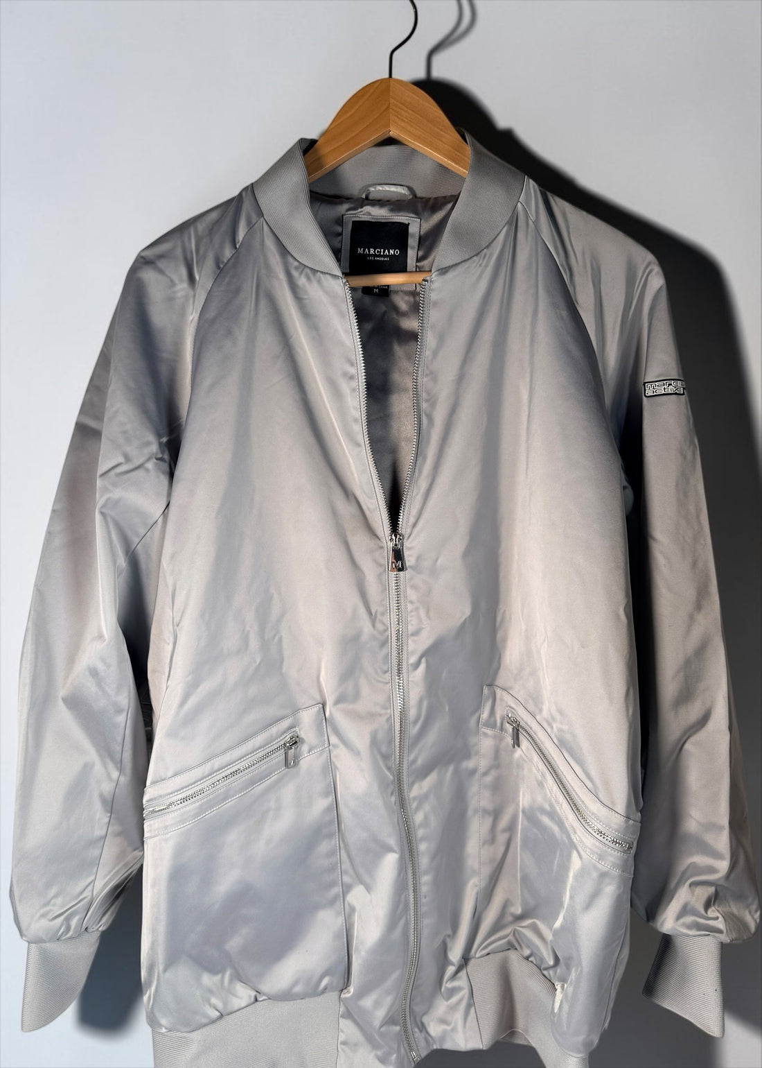 Shop the Marciano Limitless bomber jacket in silver-grey satin, size M. Original tags attached, zip pockets and ribbed trim. Minor marks disclosed.