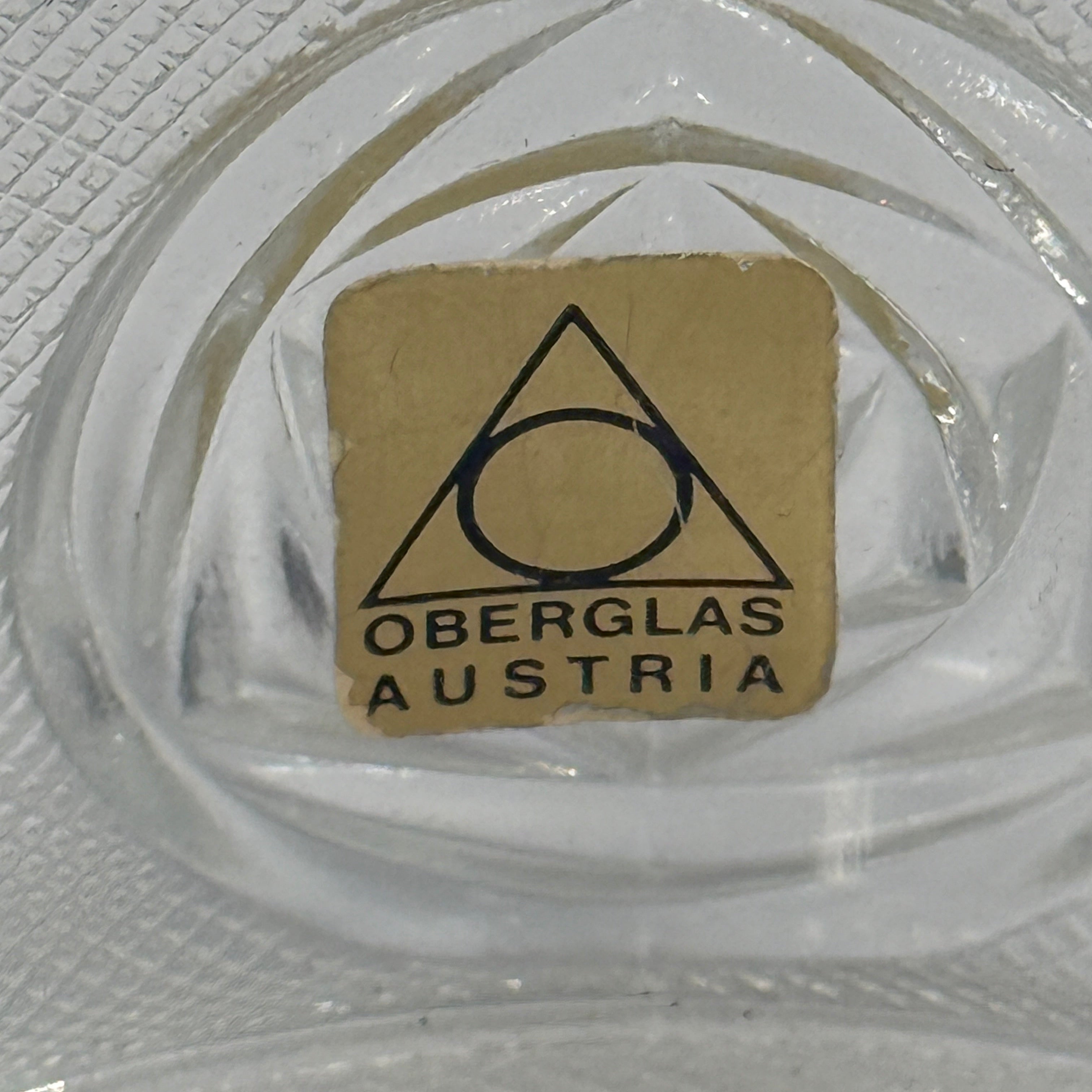 Clear glass object with a small brown label featuring 'Oberglas Austria' on a textured surface.