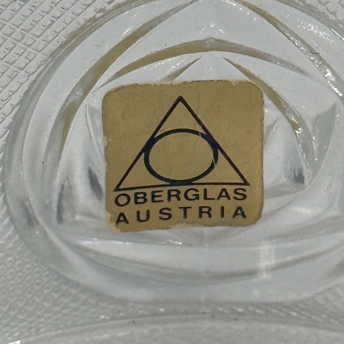 Clear glass object with a small brown label featuring 'Oberglas Austria' on a textured surface.