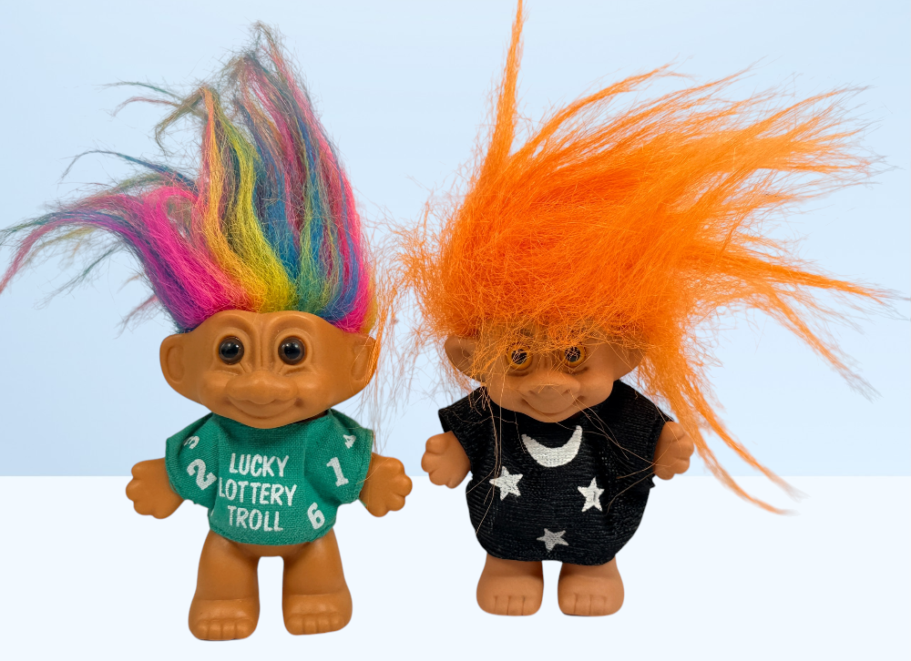 Two small troll figurines standing side by side against a light blue and white background. The left troll has multicolored hair in pink, yellow, green, and blue, wearing a green shirt that reads 'LUCKY LOTTERY TROLL'.
