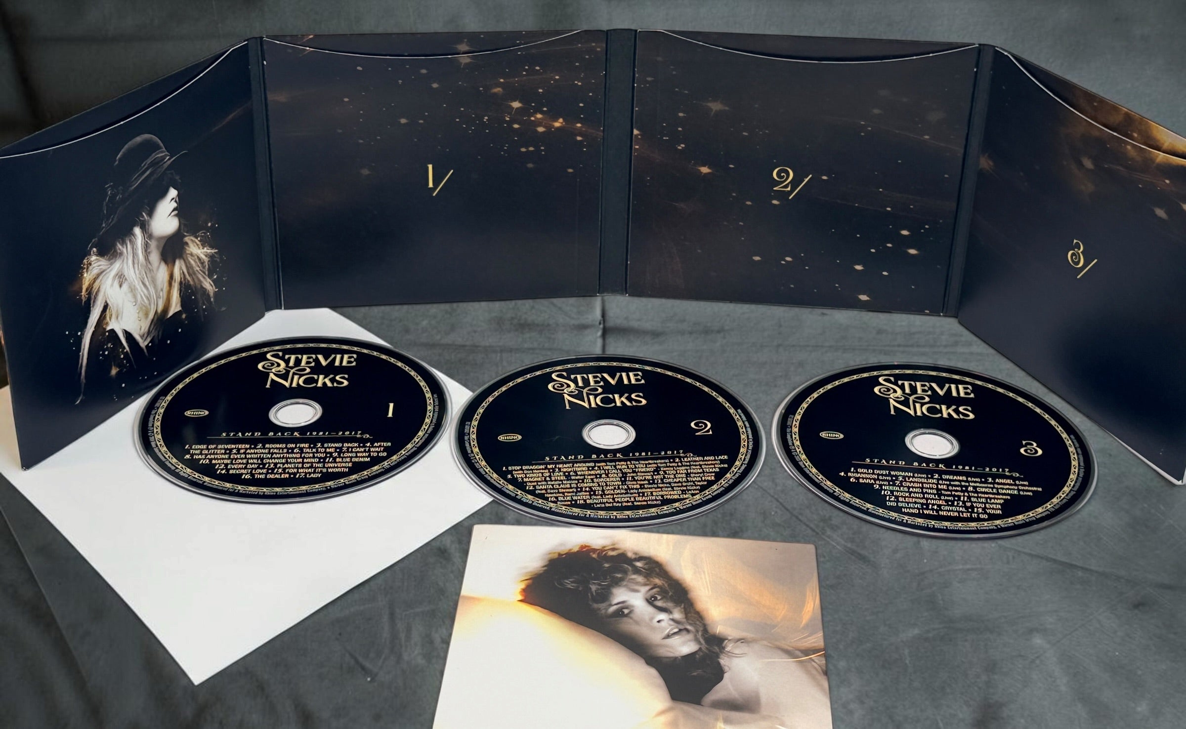 A Stevie Nicks 'Exotica' 3-disc CD box set displayed open on a dark gray surface. The set includes three black CDs with gold lettering, a black fold-out cover with a portrait of Stevie Nicks, a white insert page, and a small printed photo card.