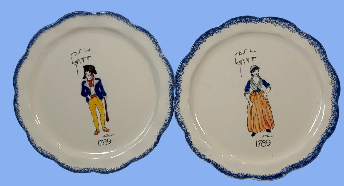 Two ceramic plates with decorative blue rims and illustrations of people from 1789 against a white brick wall.