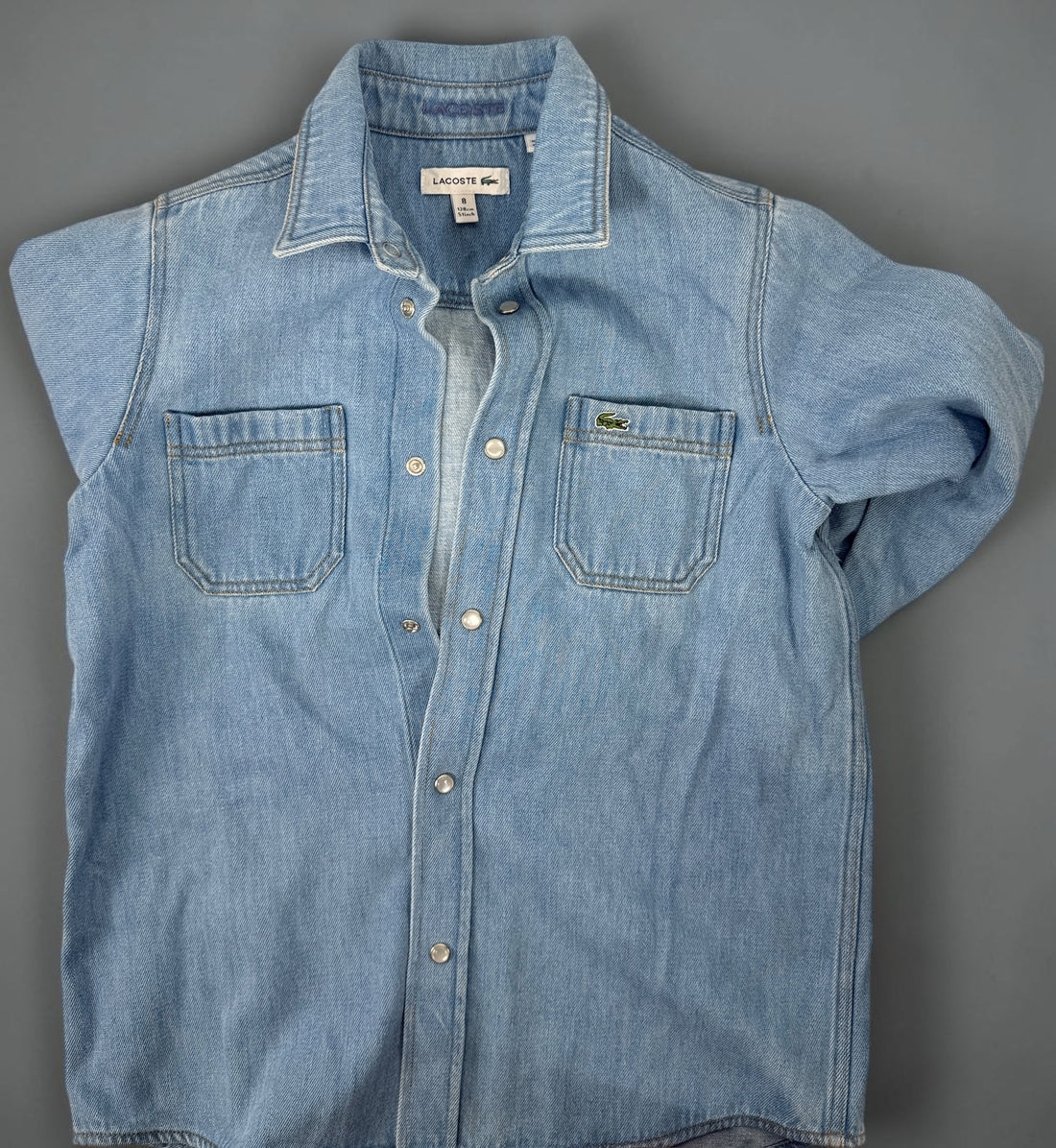 Authentic Lacoste kids denim overshirt in size 8 featuring snap-button closure, chest pockets, and classic crocodile logo. Light wash designer children’s wear.
