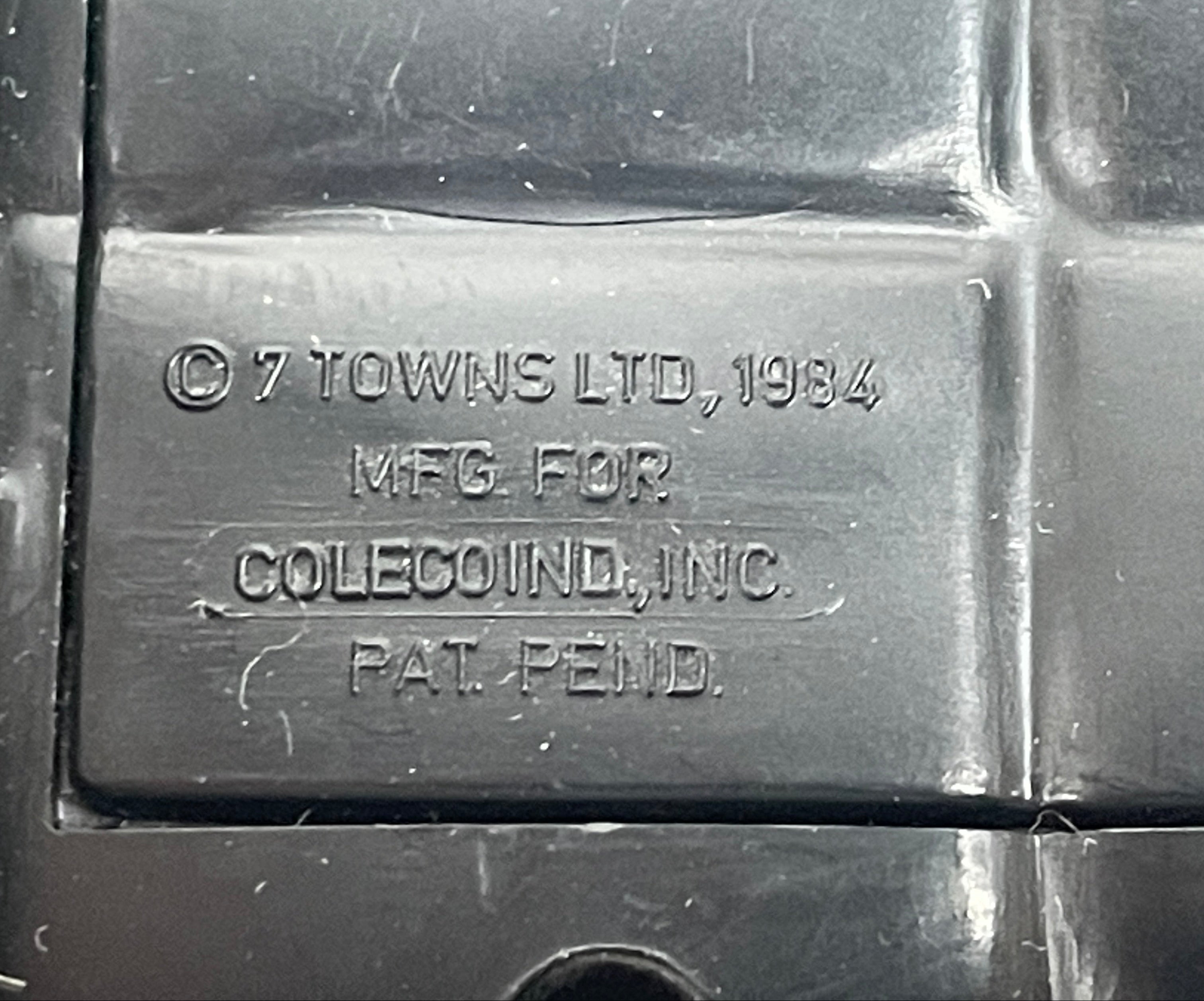 Metal plaque with '7 Towns Ltd, 1984' and 'MFG FOR COLECOIND, INC.' text on a textured surface.