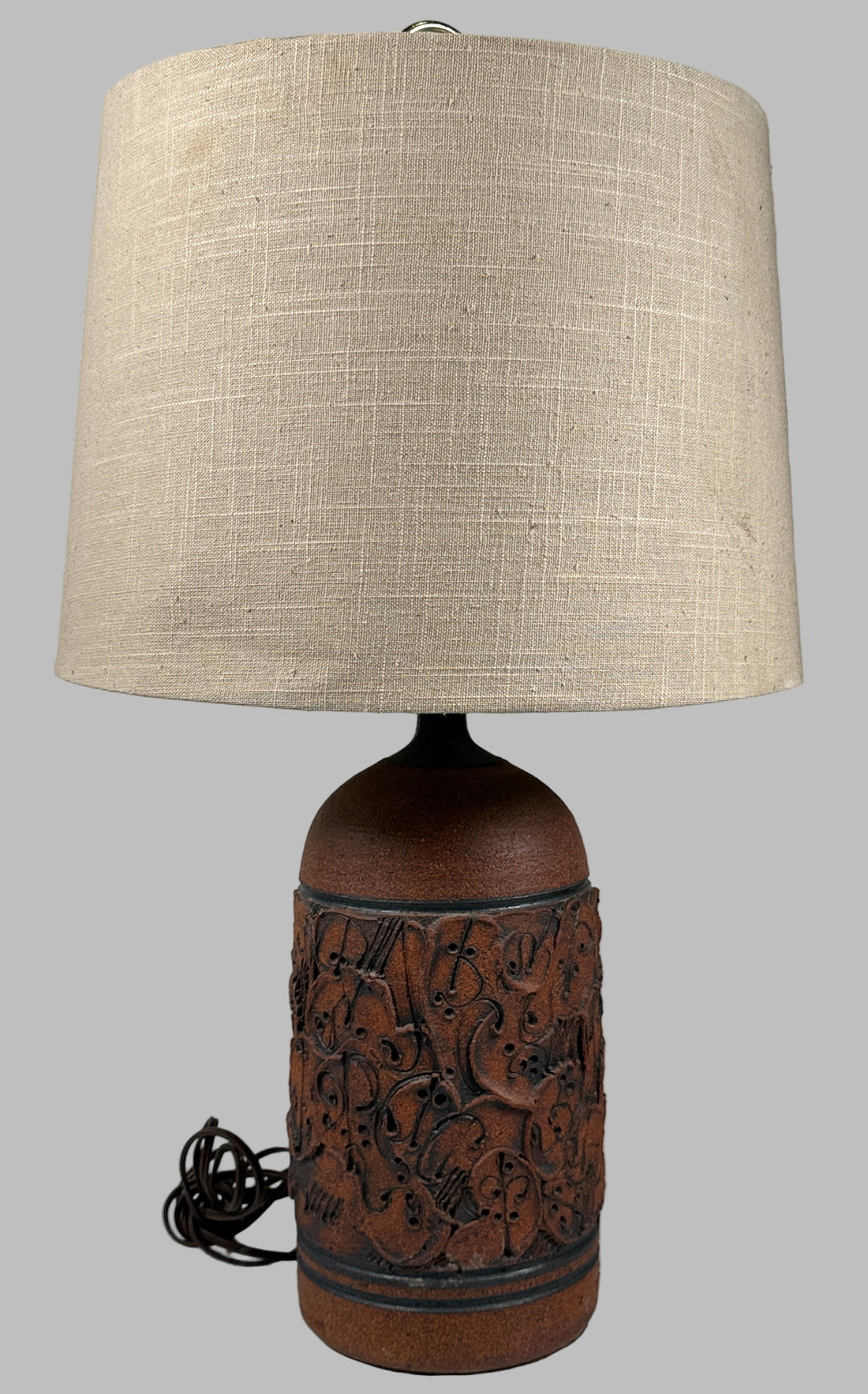 Table lamp with a textured brown base and beige lampshade against a gray brick wall.