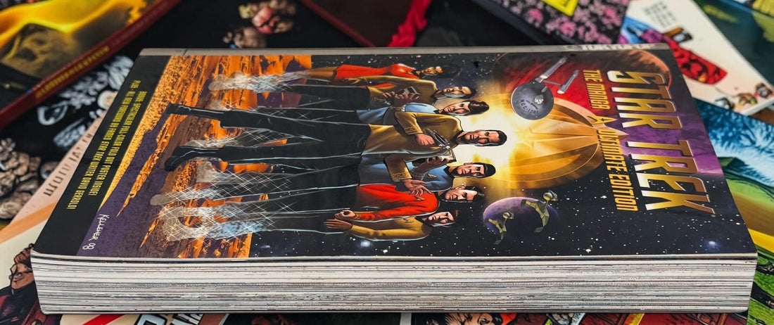 A hardcover book or comic-style publication titled Star Trek: The Official Movie Edition, shown standing upright on a surface with other colorful items around it. The cover features the Star Trek logo and artwork of crew members in space-themed scenes.